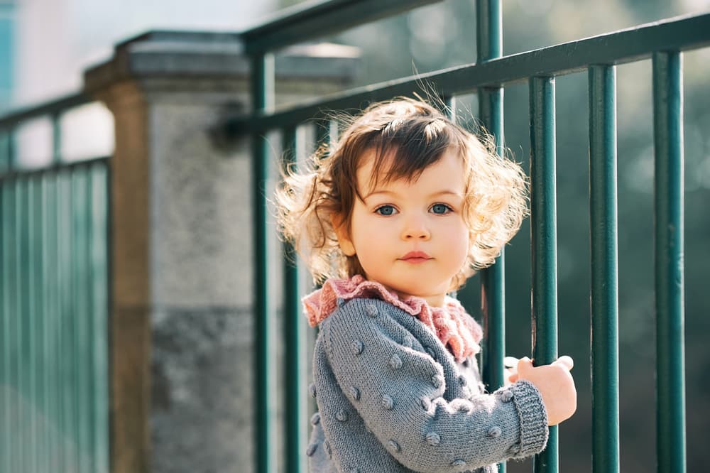 toddler girl standing at iron fence with gray sweater on