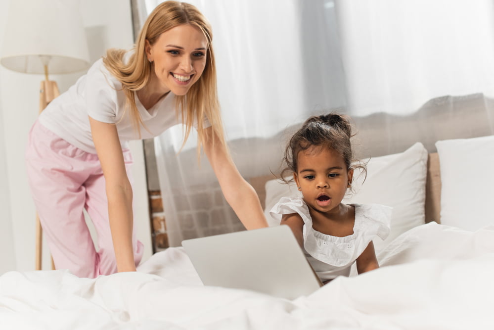 toddler girl on bed with laptop and mother hovering over watching