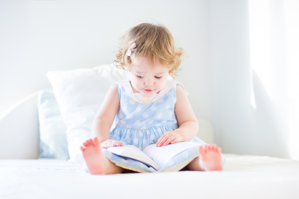 toddler girl sitting on bed reading a book